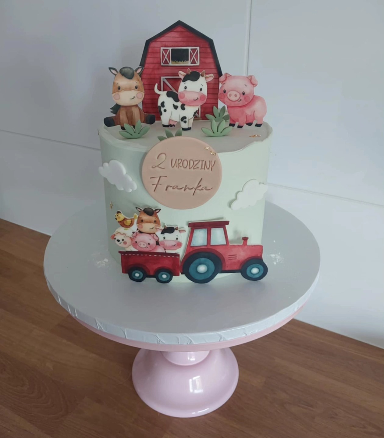 Tort na 2 urodziny Franka, zdobiony motywami farmy: zwierzęta, stodoła, traktor. Pastelowa kolorystyka, tort na różowej paterze.