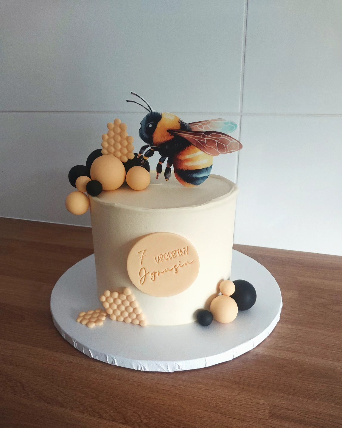 Tort urodzinowy z motywem pszczoły, dekorowany kulkami i plastrami miodu, z napisem '7 Urodziny Jędrusia'. Kremowy tort na drewnianym blacie.