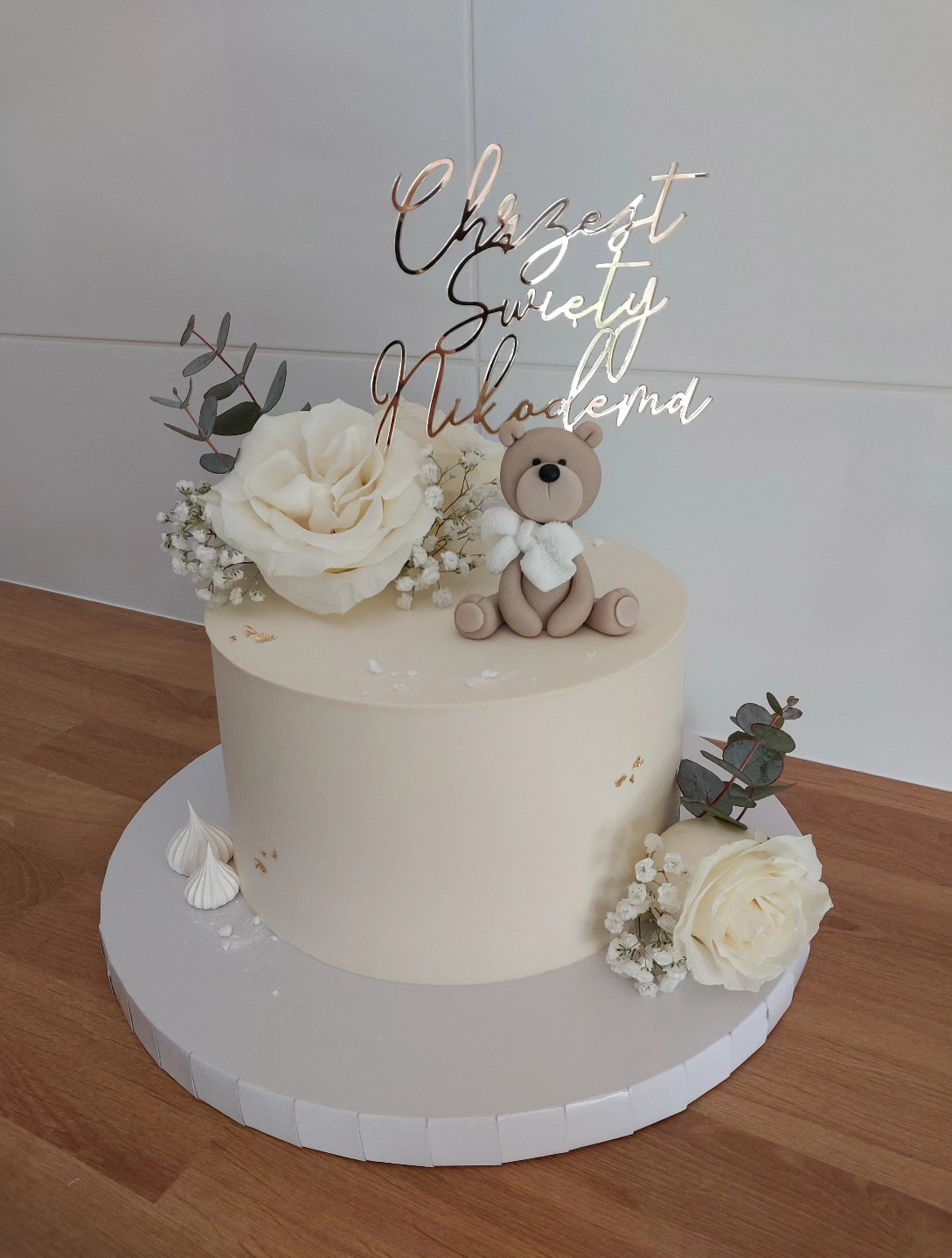 Tort na chrzest z misiem, białymi różami i napisem 'Chrzest Święty Nikodema' w złotym kolorze. Minimalistyczny design i delikatne zdobienia.