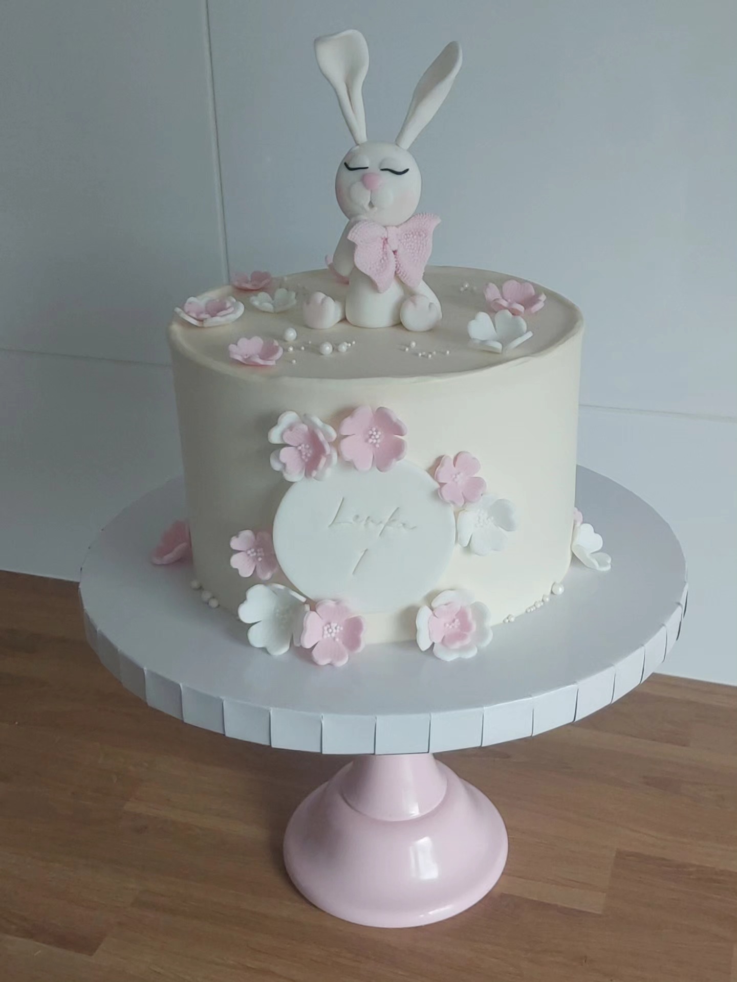 Tort urodzinowy z kremem, dekorowany kwiatami i figurką królika z zamkniętymi oczami, na różowej podstawie. Dekoracja z napisem 'Lenka 7'.