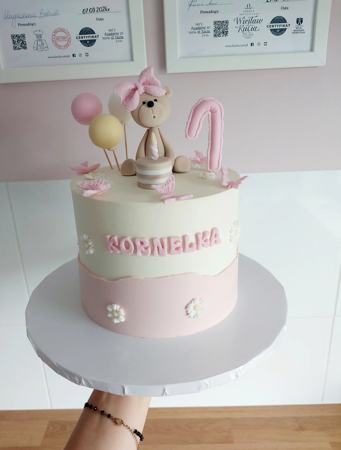 Tort urodzinowy dla Kornelki z misiem, balonami i cyfrą 1, trzymany na podeście. W tle certyfikaty. Pastelowa kolorystyka, kremowo-różowe dekoracje.