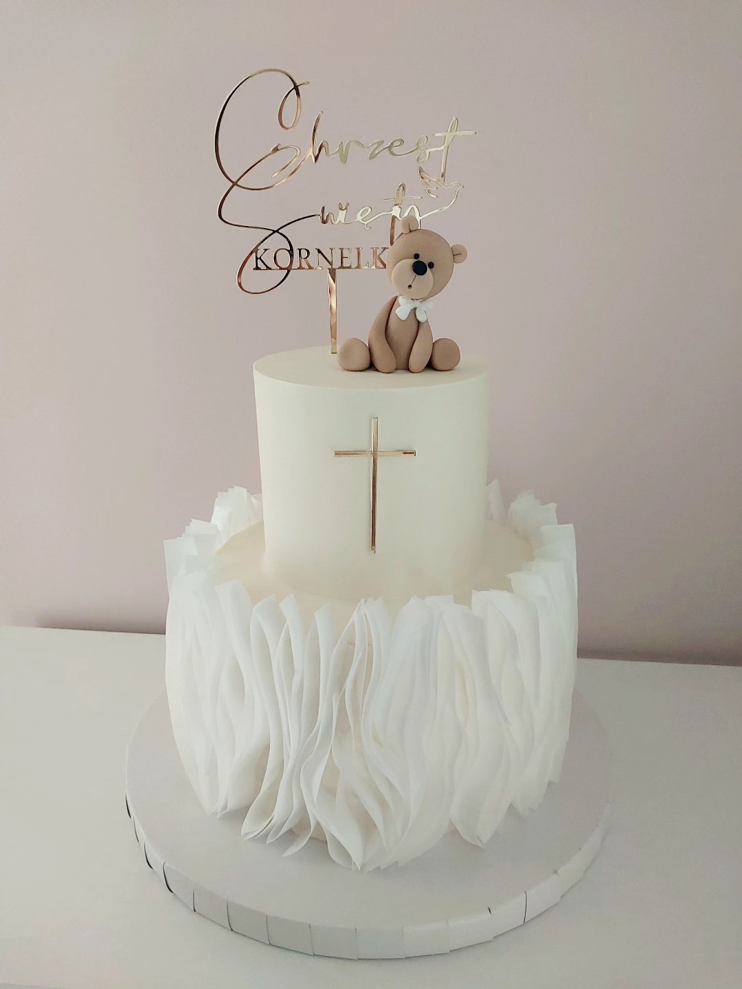 Dwupoziomowy tort w bieli, dekorowany falbanami i złotym krzyżem, z topperem 'Chrzest Święty Kornelka' i figurką misia na górze. Idealny na uroczystość.
