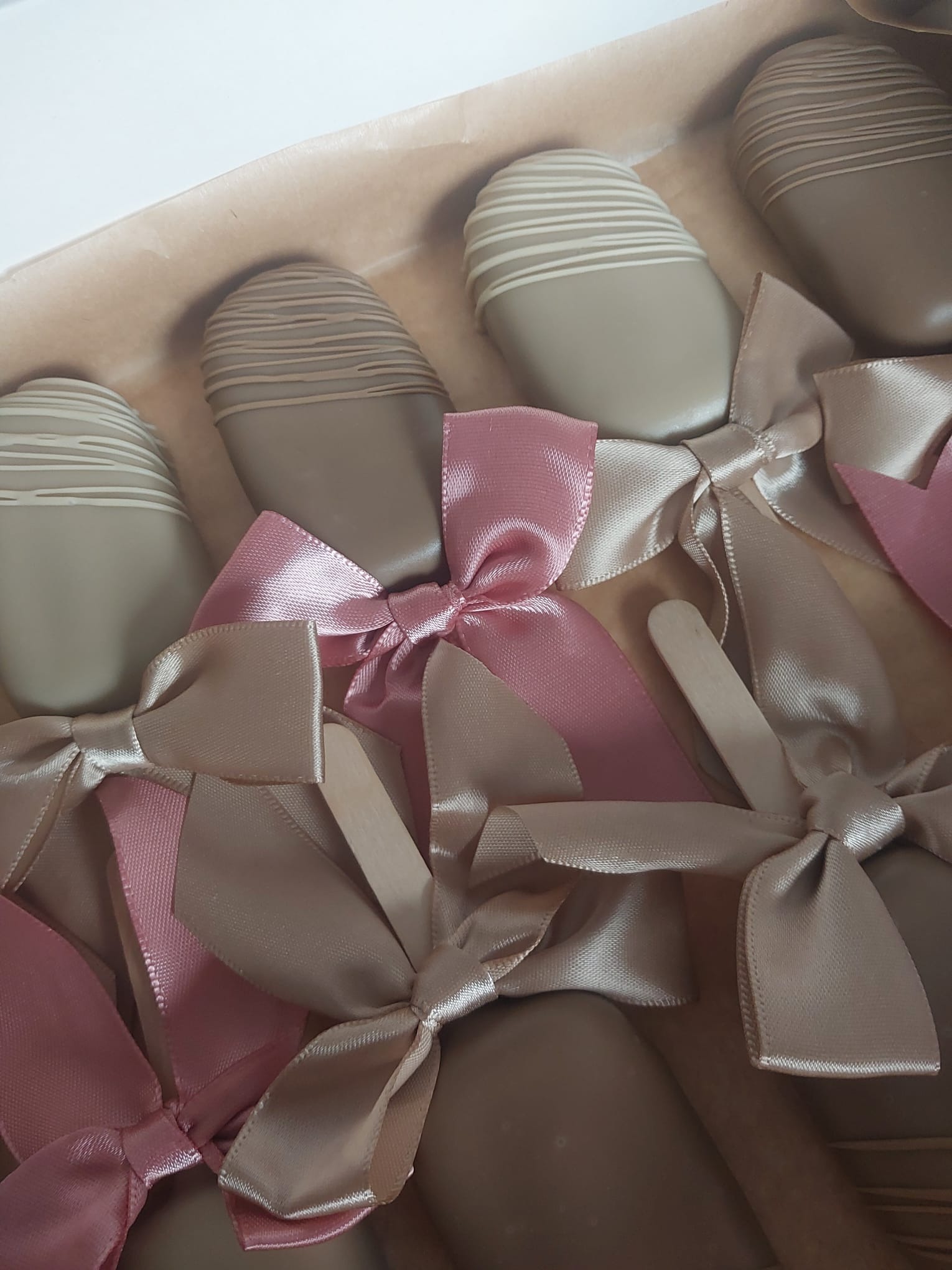 Pudełko z deserami na patyku, oblane czekoladą i ozdobione wstążkami w odcieniach różu i beżu. Delikatne paski na czekoladzie dodają elegancji.