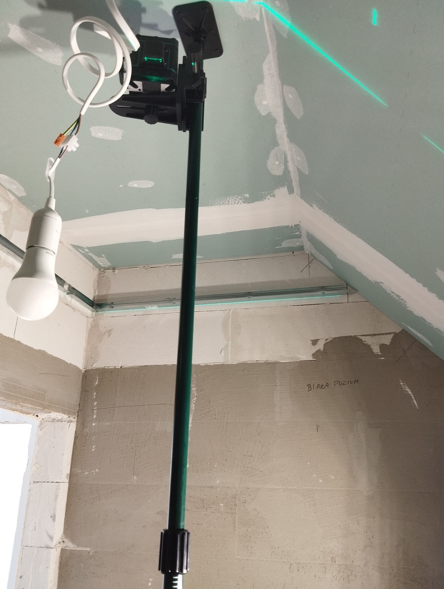 Montaż sufitu podwieszanego z płyt karton-gips, widoczny laser krzyżowy wyznaczający poziom, oświetlenie robocze i fragment ściany z napisem 'Biała Puzium'.
