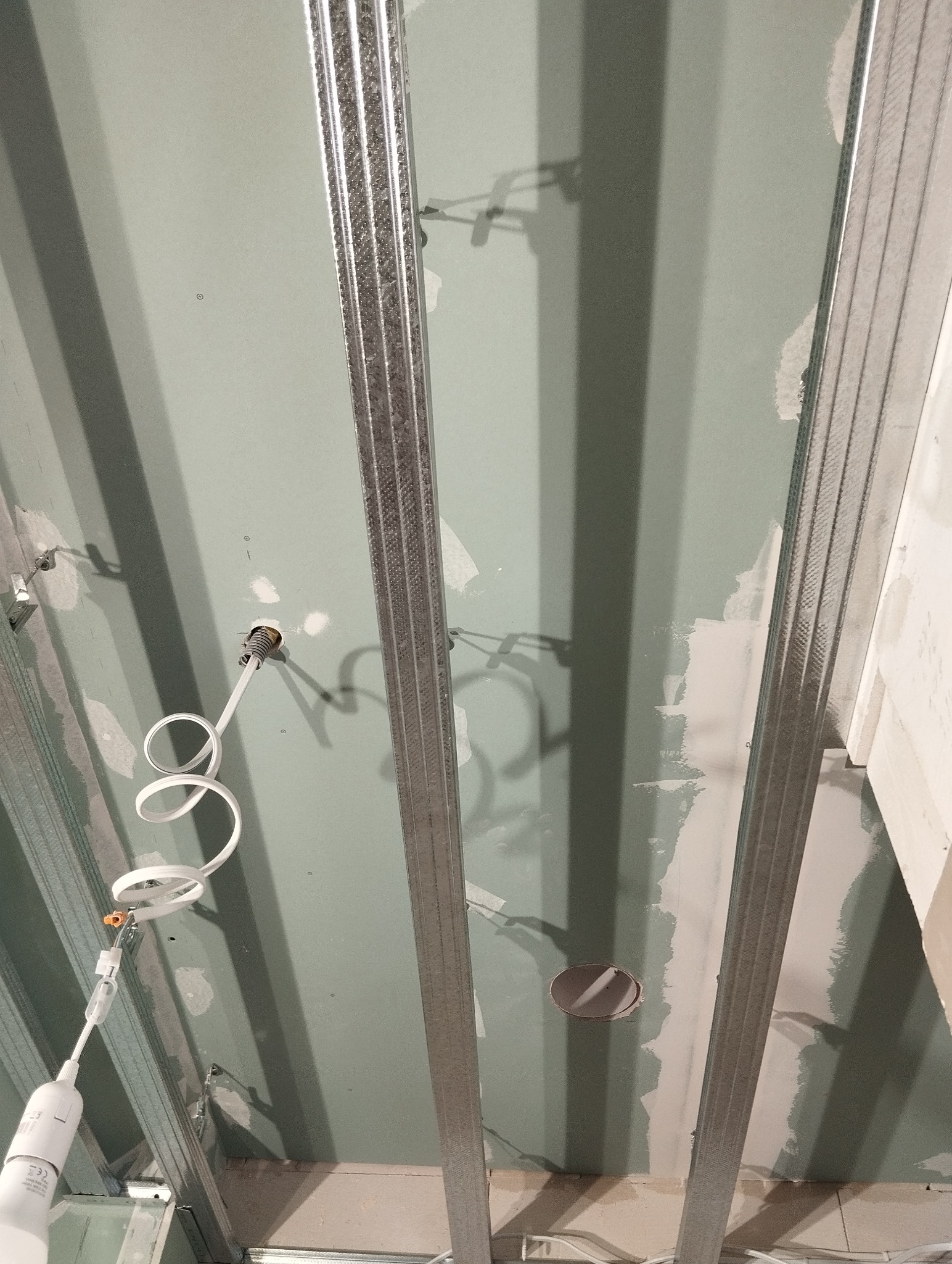 Montaż sufitu z płyt karton-gips: widoczne profile, płyty z otworami na oświetlenie i prowizoryczne oświetlenie budowlane. Etap prac wykończeniowych.