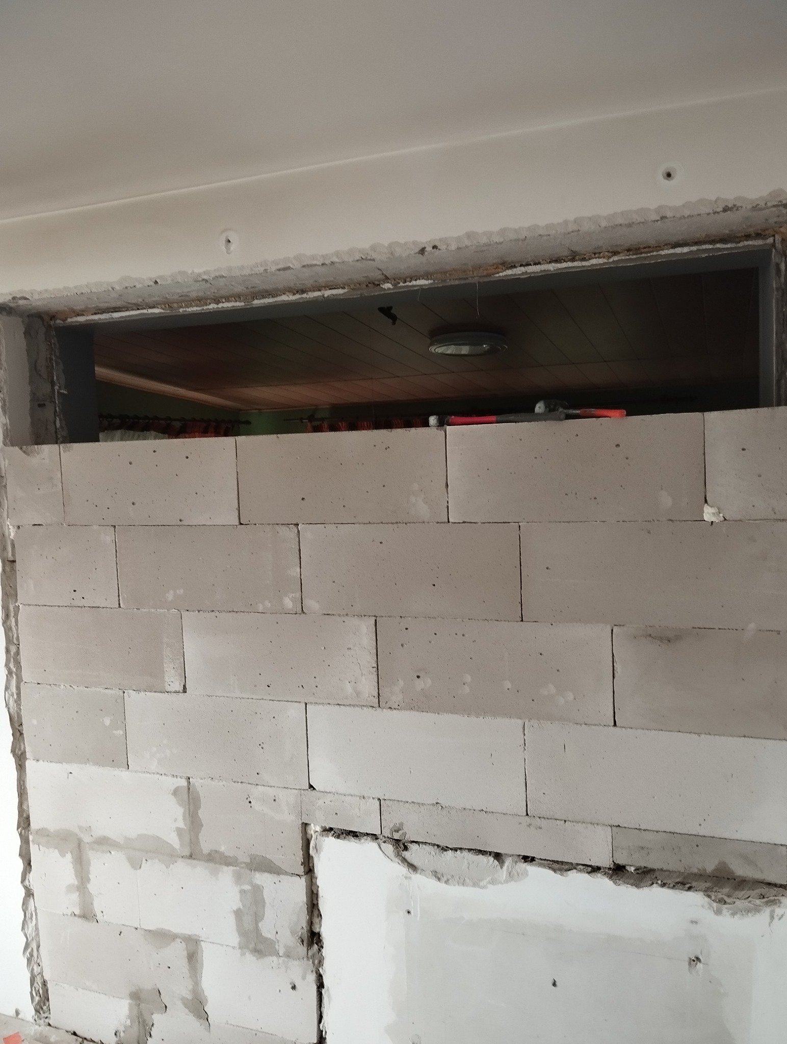 Ściana z białych bloczków, widoczna wnęka okienna z fragmentem sufitu i lampy. Częściowo usunięty fragment ściany gipsowo-kartonowej. Etap prac murarskich wewnątrz budynku.