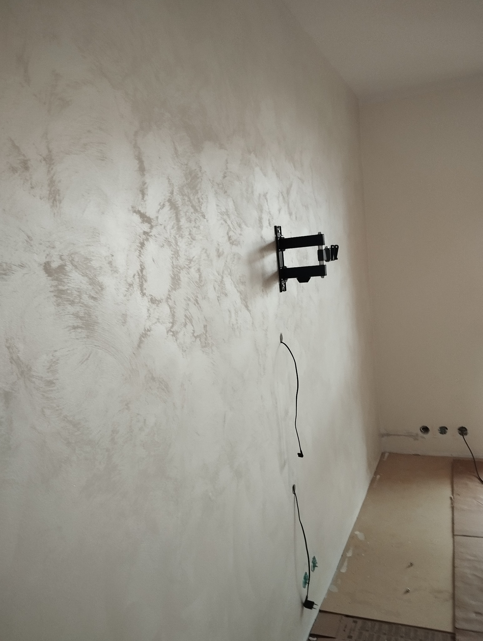 Ściana z dekoracyjnym tynkiem w odcieniach bieli i szarości, widoczny uchwyt na telewizor i wystające przewody. Podłoga zabezpieczona kartonem.