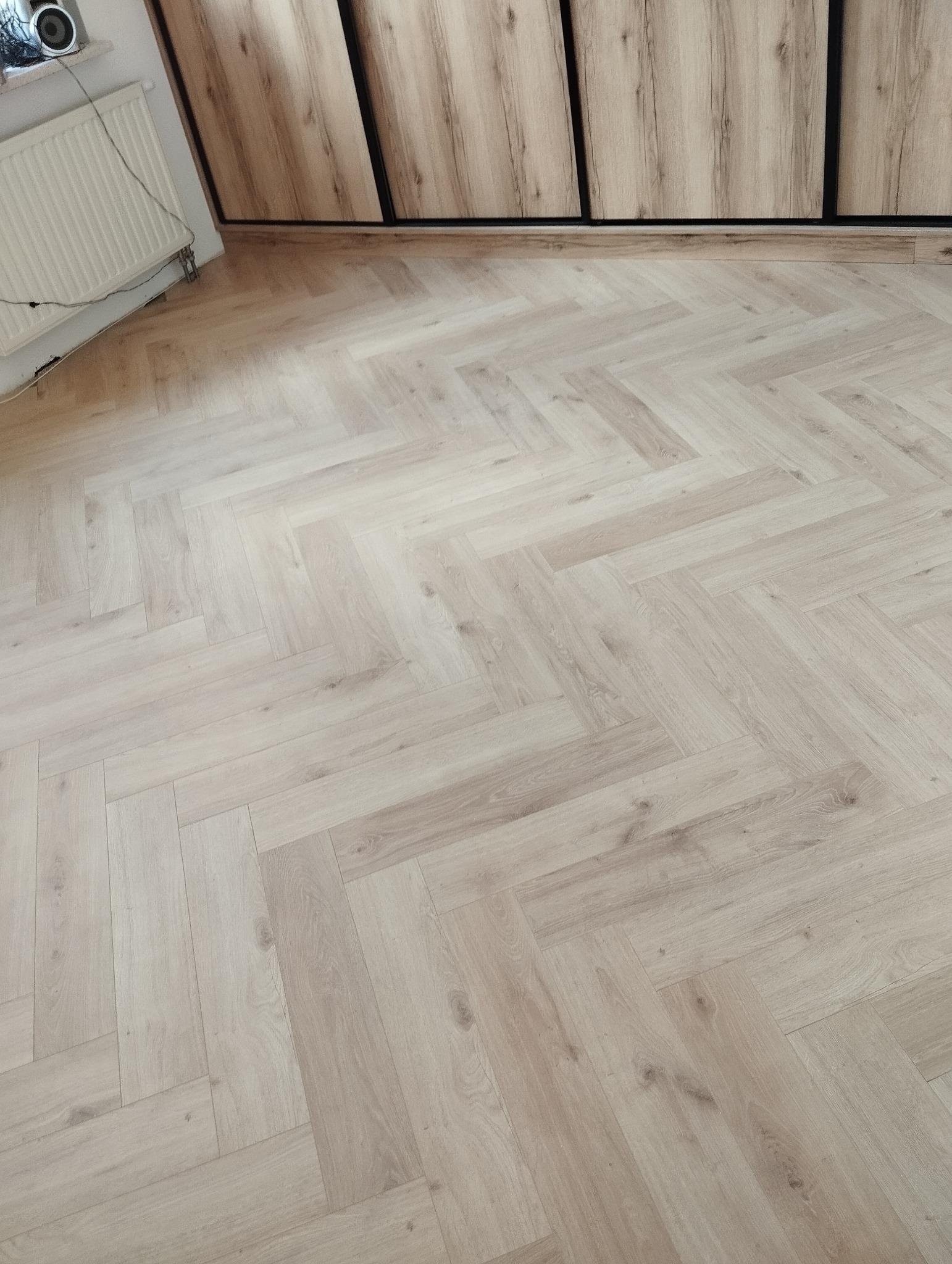 Jasna, drewniana podłoga ułożona w jodełkę, widok z góry. Na ścianie grzejnik i szafa w odcieniach drewna. Ukończona realizacja.