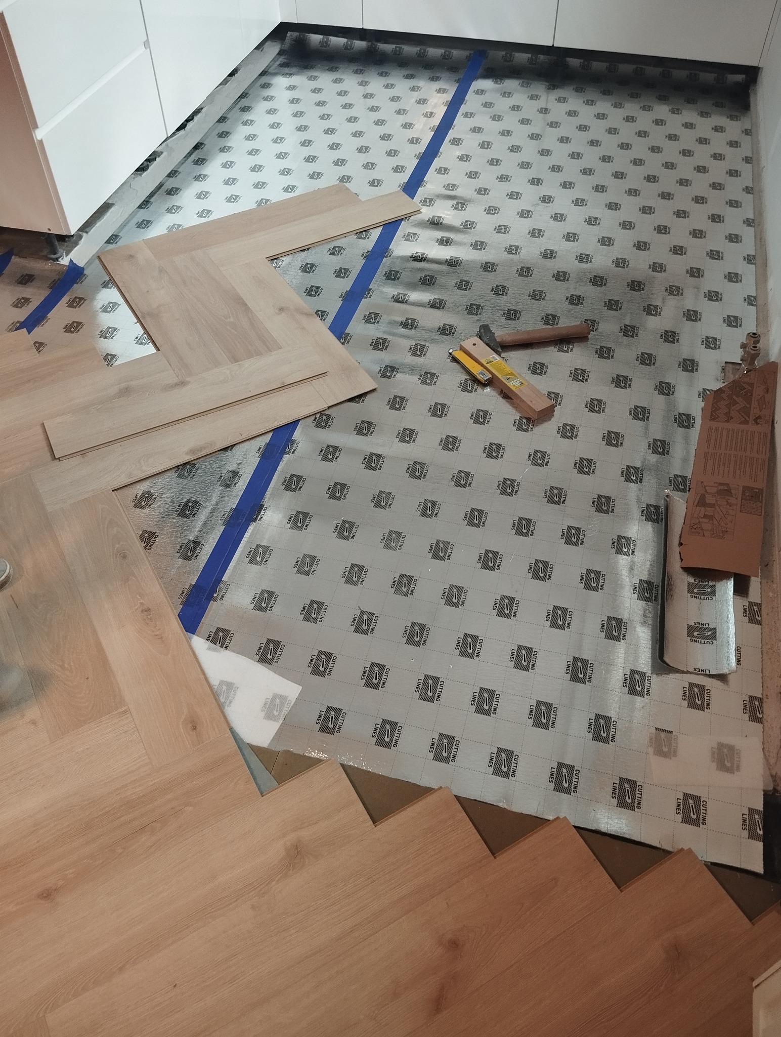 Układanie paneli podłogowych w jodełkę. Widoczne narzędzia pracy i podkład pod panele z niebieską taśmą. Fragment białej zabudowy kuchennej w tle.