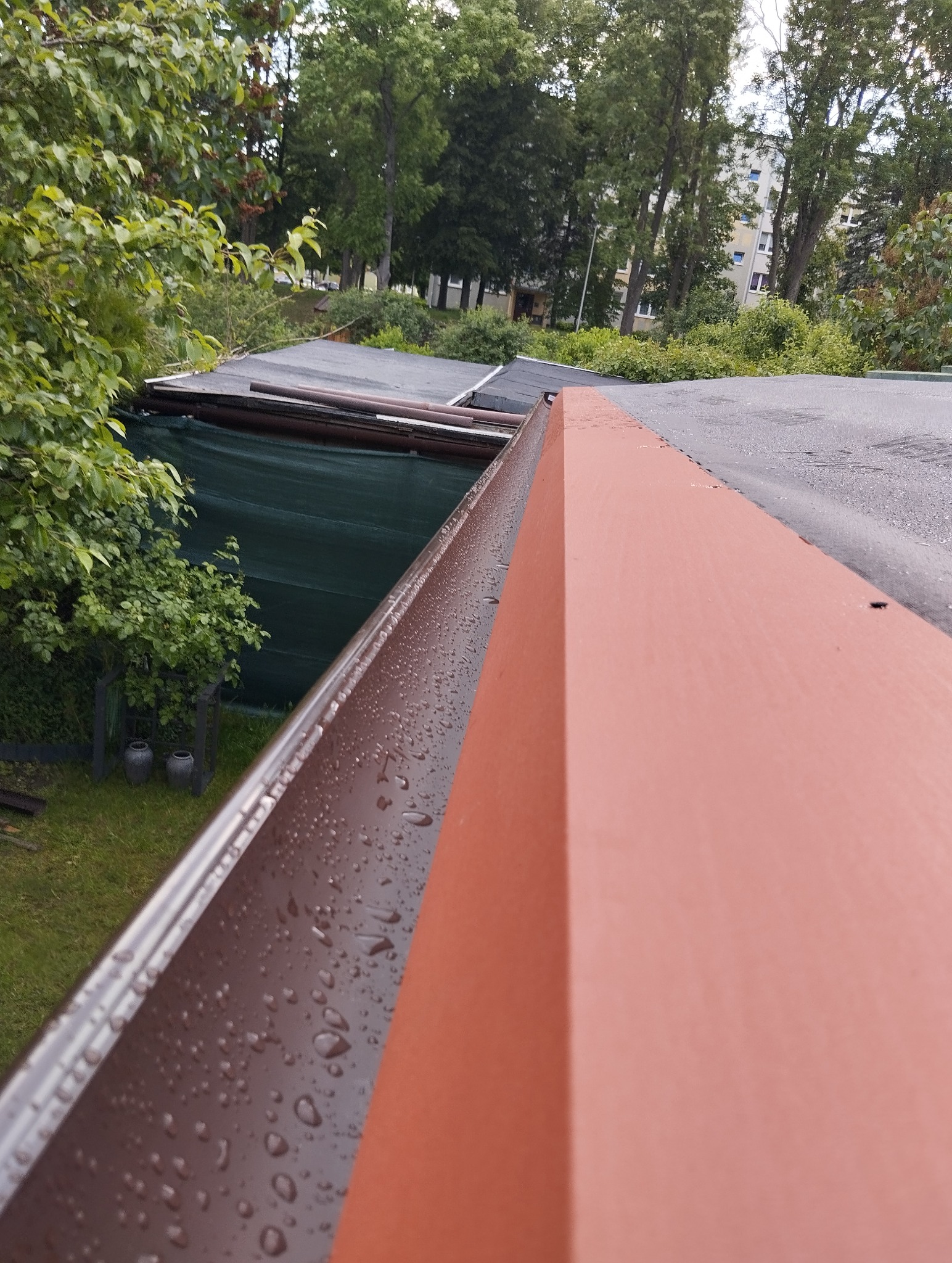 Metalowy system rynnowy w kolorze brązowym, pokryty kroplami wody, z bliska. Widok z góry na dach budynku i zieleń w tle.