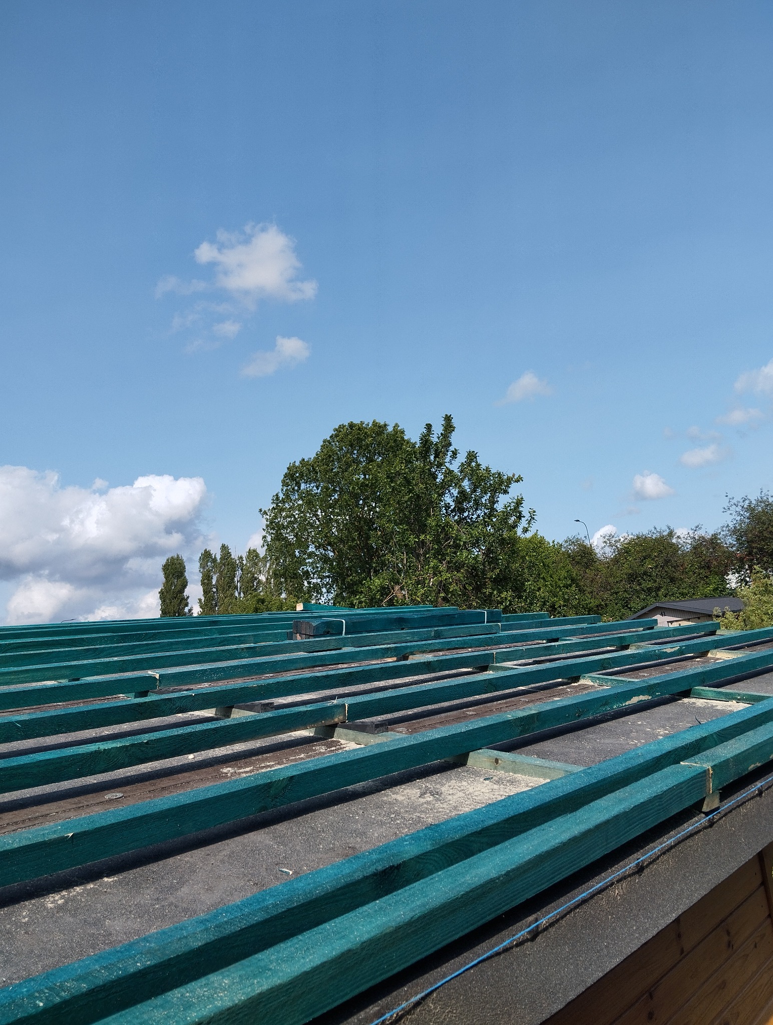 Szkielet dachu z zielonych belek na czarnej membranie, widok z dołu na błękitne niebo z chmurami i drzewa w tle. Konstrukcja dachu w trakcie budowy.