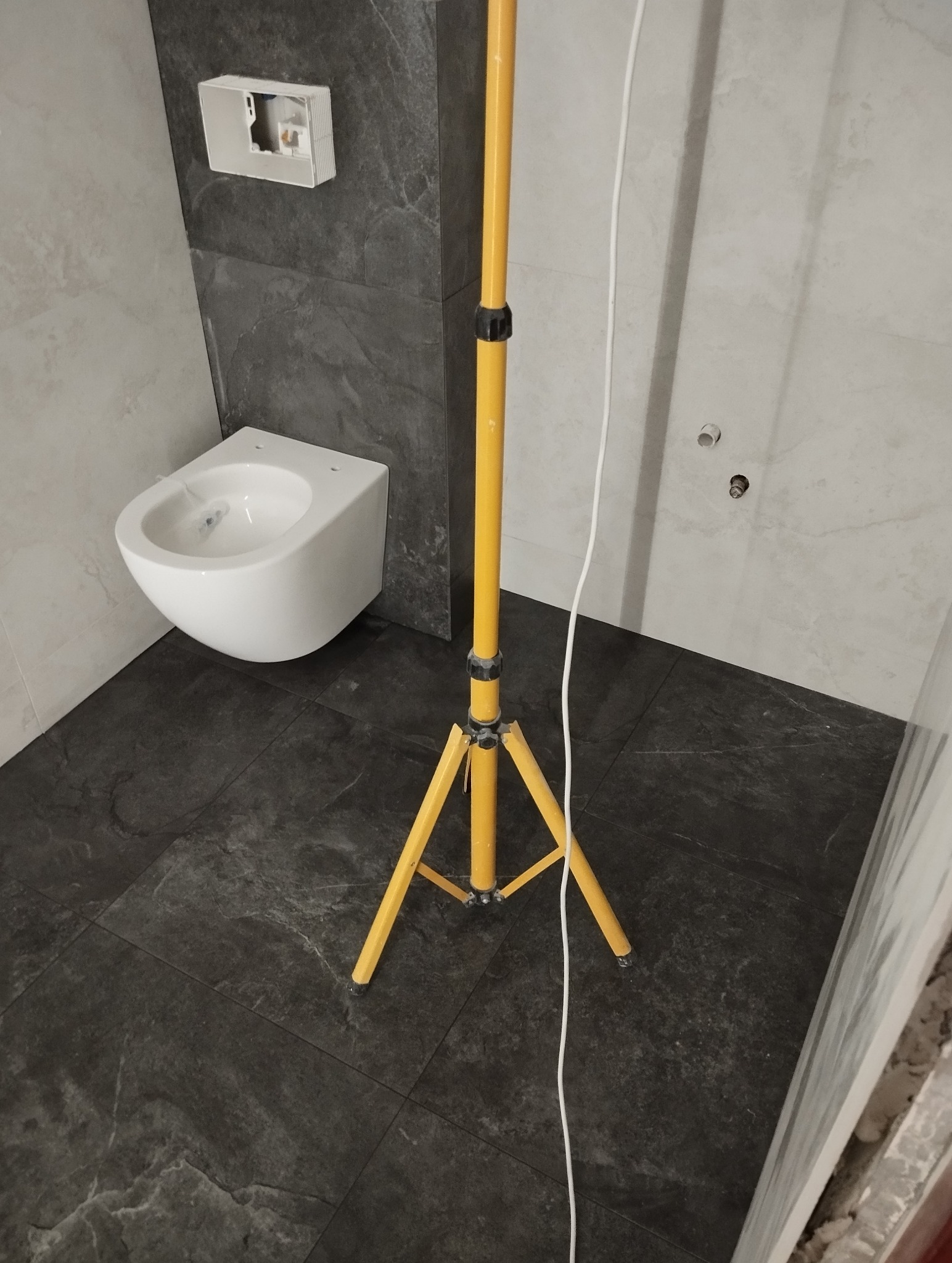 Nowoczesna, częściowo wykończona łazienka z podwieszaną toaletą, ciemną podłogą i żółtym statywem oświetleniowym. Widoczna instalacja.