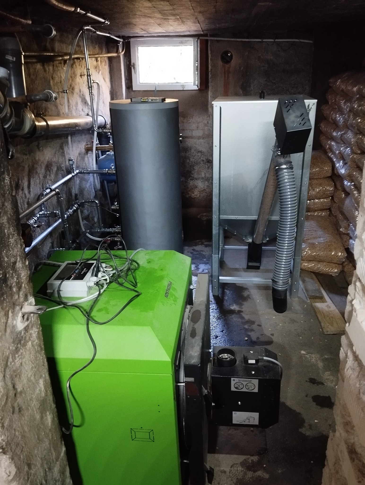 Kotłownia z nowoczesnym piecem na pellet, zbiornikiem buforowym i podajnikiem. Widoczne rury instalacji CO oraz zapas pelletu w workach. Oświetlenie sufitowe.