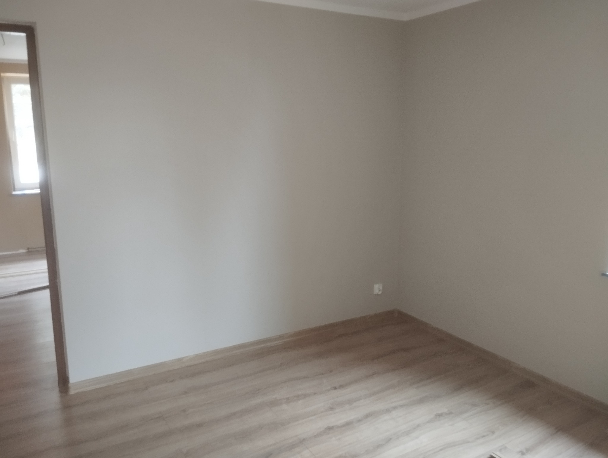 Jasne wnętrze po remoncie: ściany pomalowane na beżowo, ułożona podłoga z paneli imitujących drewno. Widoczny fragment okna w sąsiednim pomieszczeniu.