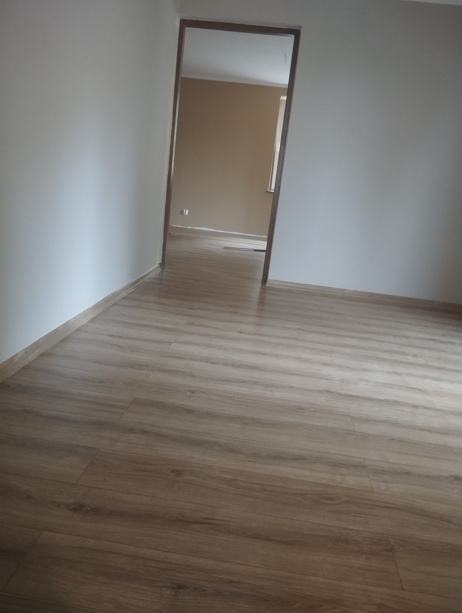 Jasne panele podłogowe w nowym pokoju, widok na sąsiednie pomieszczenie przez otwarte drzwi. Świeżo pomalowane, białe ściany, listwy przypodłogowe.