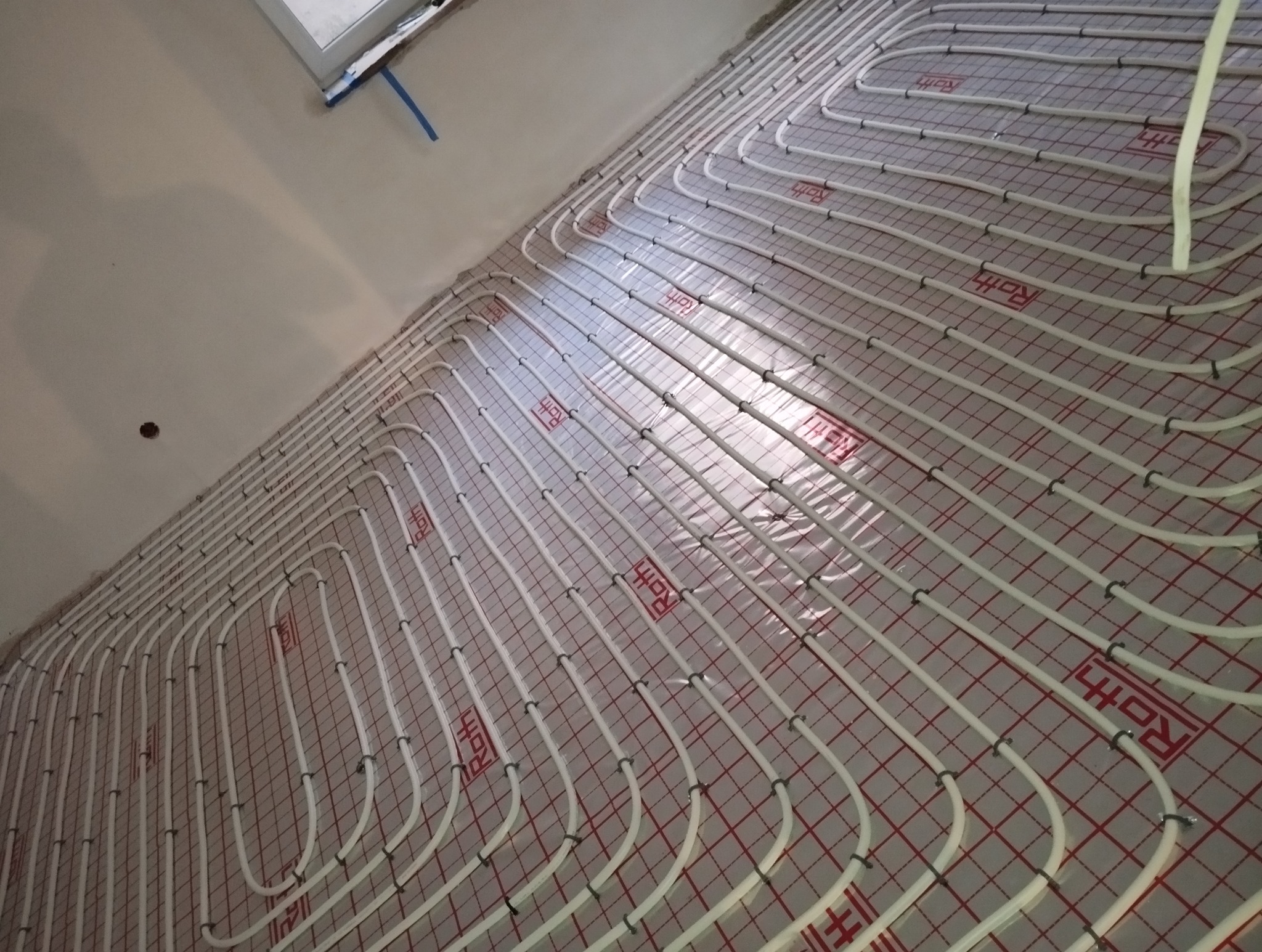 Białe rurki ogrzewania podłogowego Roth ułożone na czerwonej siatce w surowym wnętrzu, widok z góry. Instalacja przed wylewką.