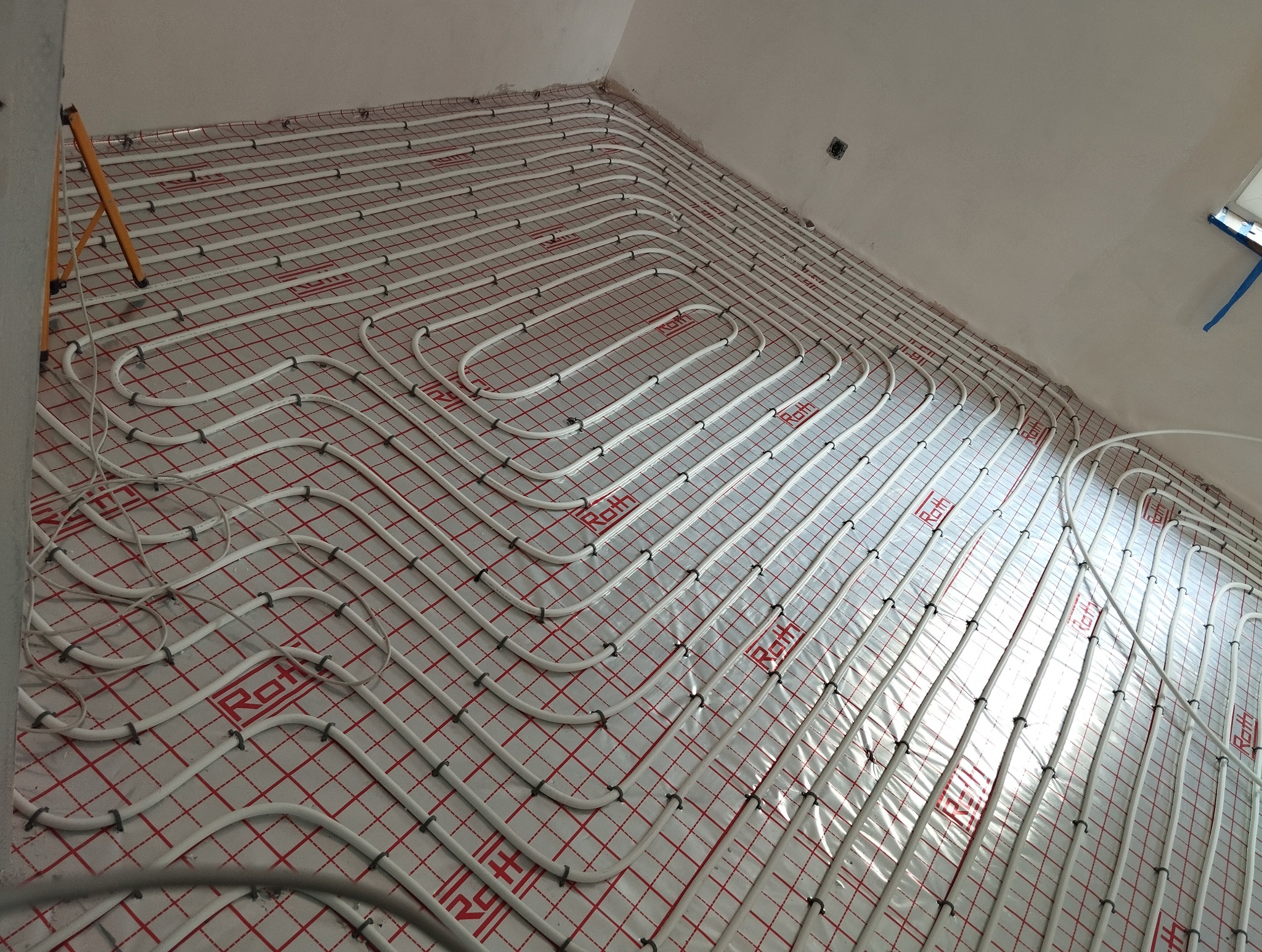 Białe rury ogrzewania podłogowego ułożone na czerwonej siatce, widok z góry, instalacja Roth, przygotowane do wylewki, etap prac wykończeniowych.
