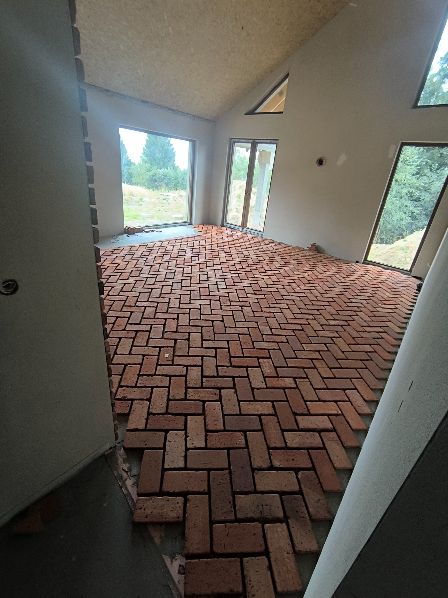 Układanie ceglanej podłogi w jodełkę w surowym wnętrzu. Widoczne okna z widokiem na zieleń. Cegły w odcieniach czerwieni i brązu tworzą regularny wzór.