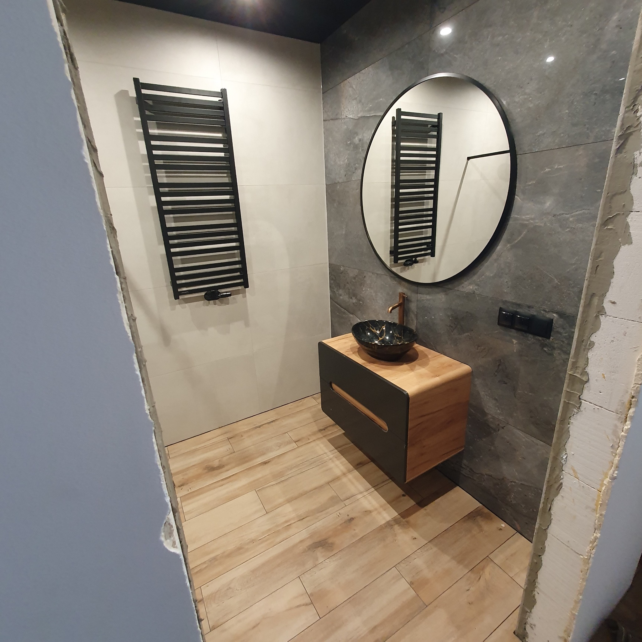 Nowoczesna łazienka po remoncie: drewnopodobne płytki, szara ściana, okrągłe lustro, szafka z umywalką i grzejnik drabinkowy. Widoczne surowe wykończenie przy futrynie.