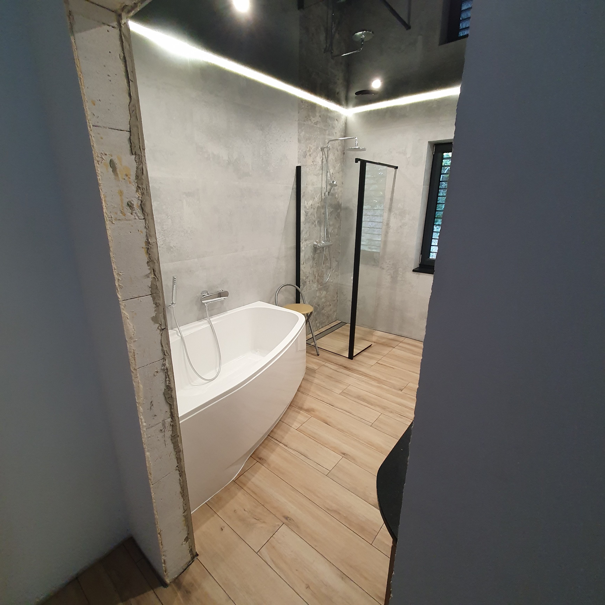 Nowoczesna łazienka z wolnostojącą wanną, kabiną prysznicową w czarnej ramie i drewnopodobną podłogą. Widok zza fragmentu ściany w trakcie prac wykończeniowych.