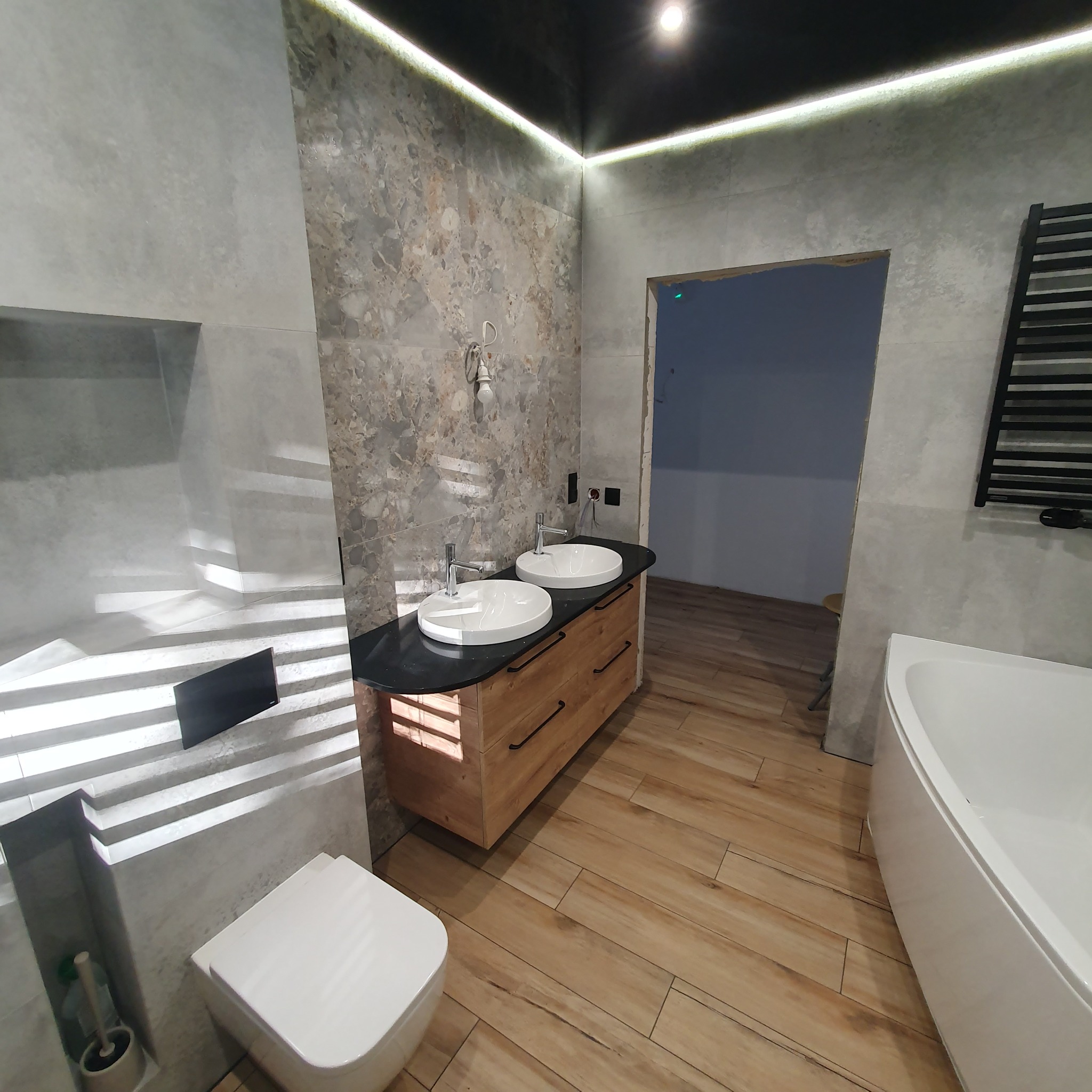 Nowoczesna łazienka z podwójną umywalką na drewnianej szafce, płytkami imitującymi kamień i oświetleniem LED. Widoczna wanna i toaleta.