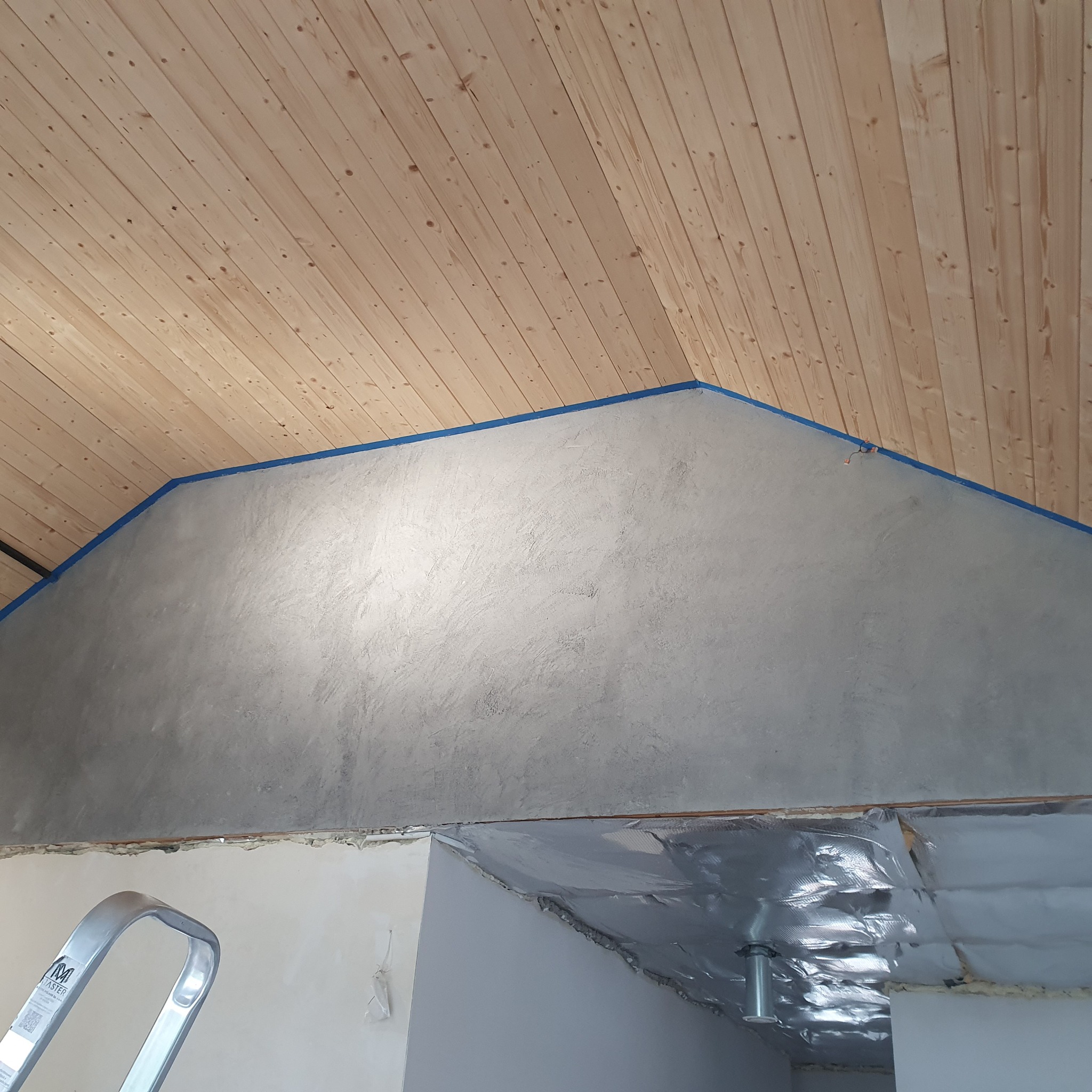 Ściana po szpachlowaniu w pokoju ze skośnym, drewnianym sufitem, zabezpieczona niebieską taśmą malarską. Widoczny fragment drabiny. Etap prac wykończeniowych.