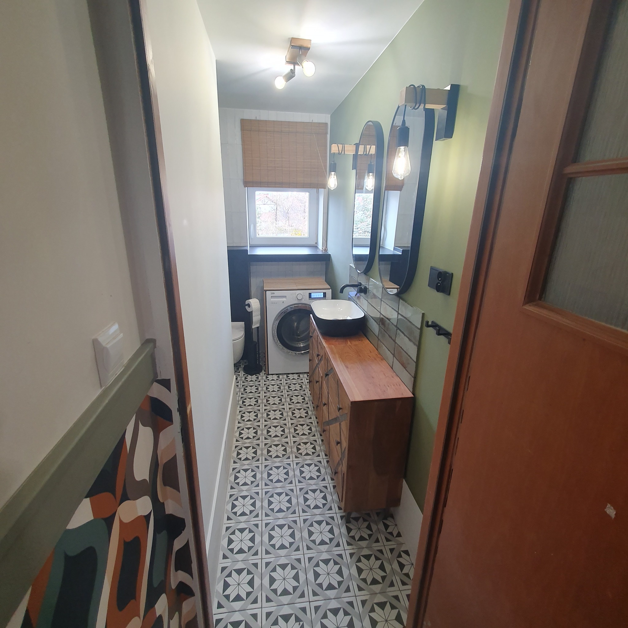 Nowoczesna łazienka z drewnianą szafką pod umywalką, wzorzystą podłogą i pralką. Widok zza drzwi, z unikalnym wzorem na ścianie po lewej stronie.