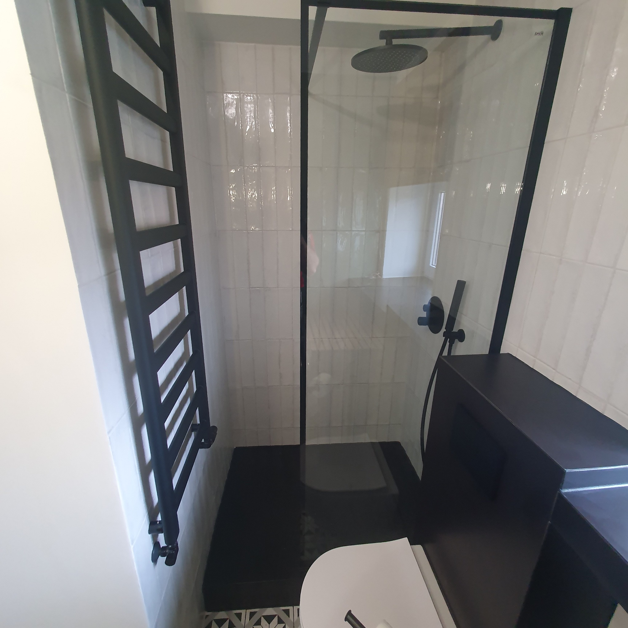 Nowoczesna łazienka po remoncie: kabina prysznicowa ze szklanymi drzwiami, czarny grzejnik drabinkowy i białe płytki na ścianach. Minimalistyczny design.