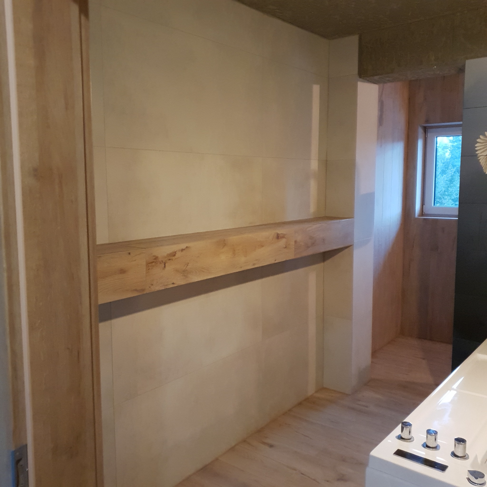 Jasna łazienka z drewnianą półką i płytkami imitującymi drewno na podłodze. Widoczne okno i fragment wanny.