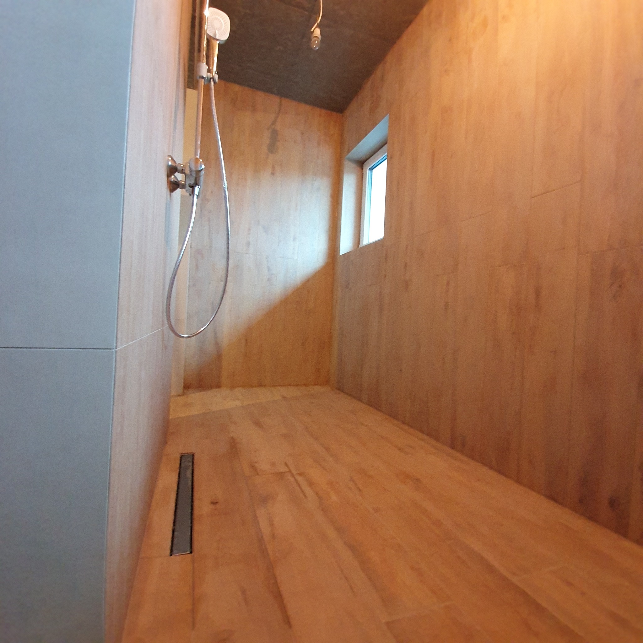 Nowoczesna kabina prysznicowa z drewnopodobnymi płytkami na ścianach i podłodze, odpływ liniowy. Minimalistyczny design, okno w tle. Widok z perspektywy.