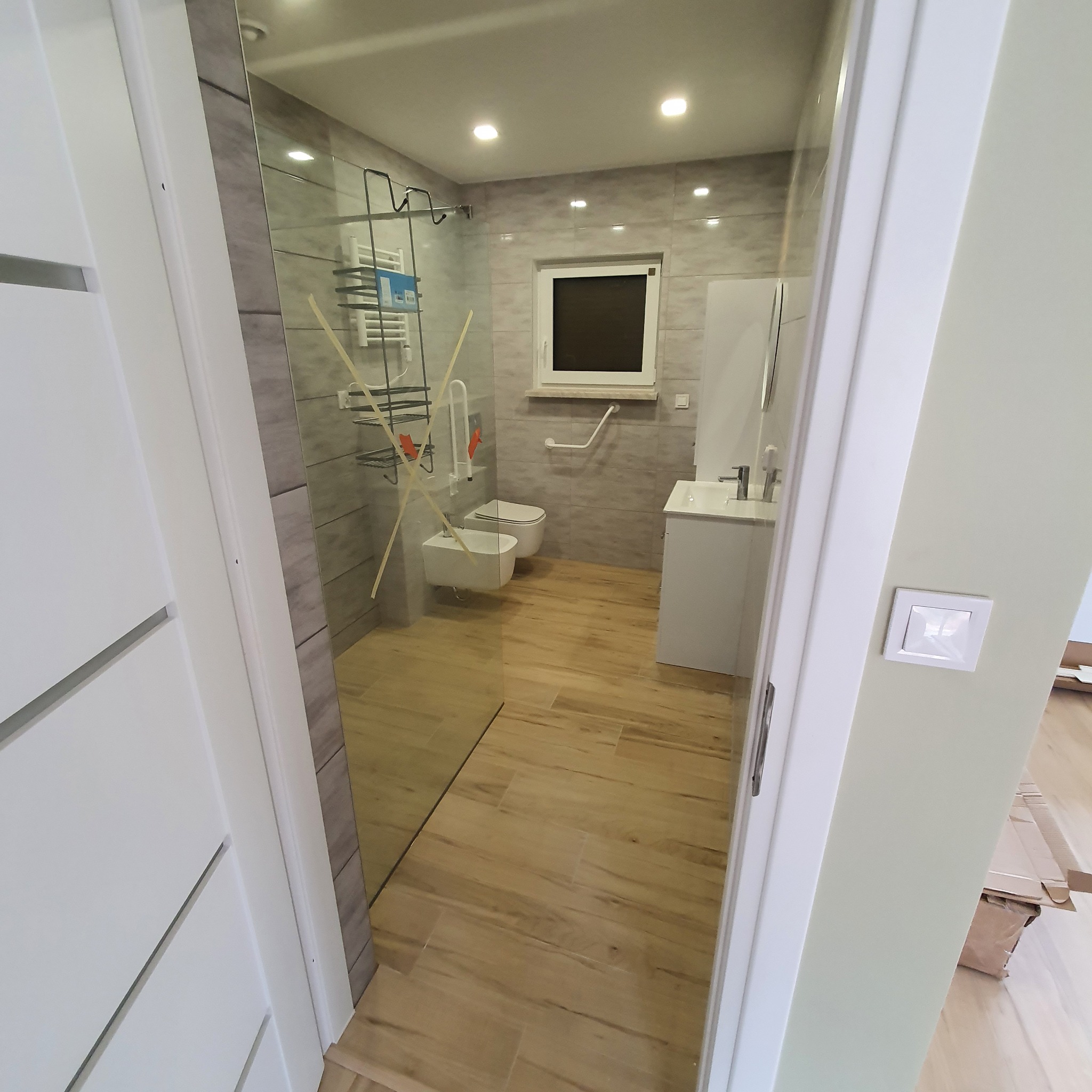 Nowoczesna łazienka z kabiną prysznicową, umywalką, toaletą i bidetem, wykończona w szaro-białej kolorystyce z drewnopodobnymi płytkami na podłodze. Widok zza uchylonych drzwi.