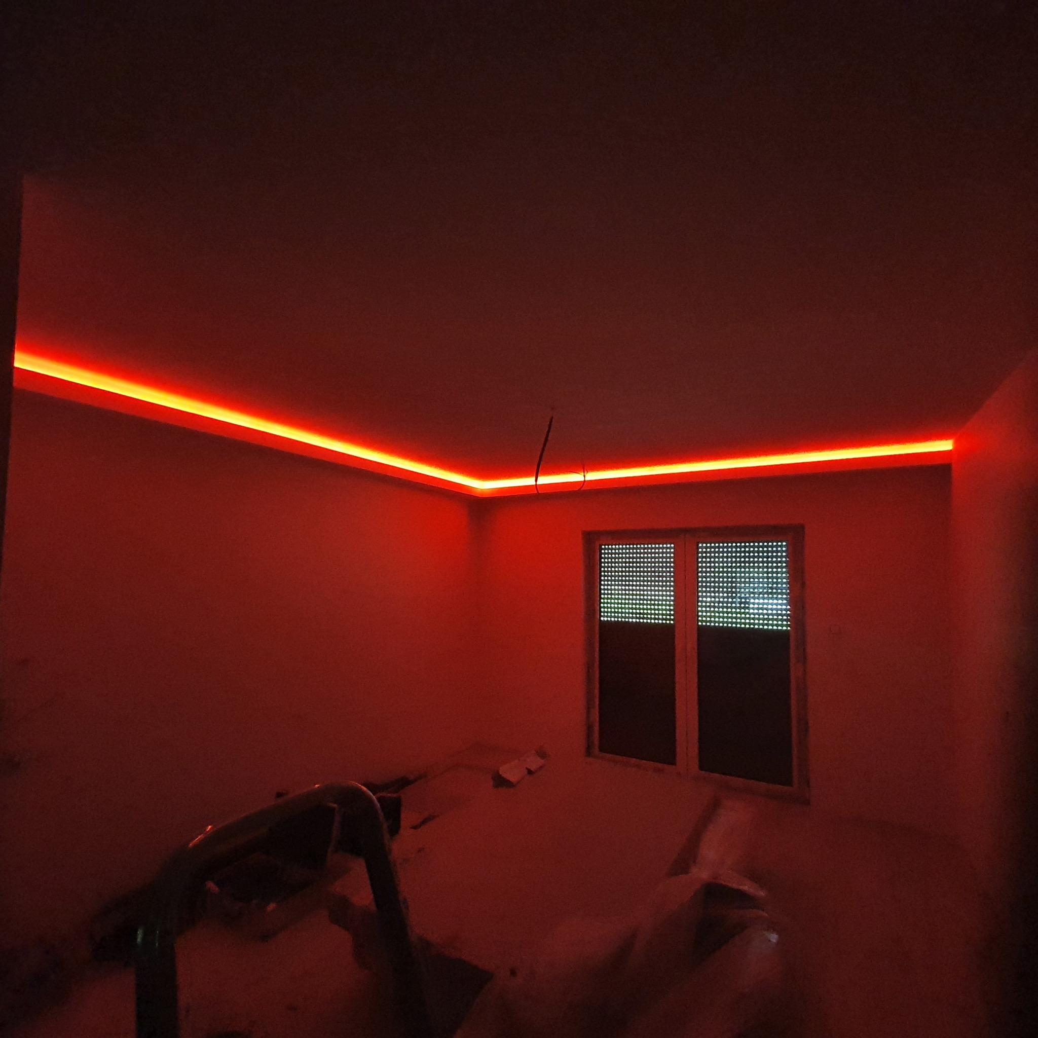 Pokój z oświetleniem LED na suficie, świecący intensywnym czerwonym światłem. Widoczne okno i elementy wyposażenia w surowym stanie.