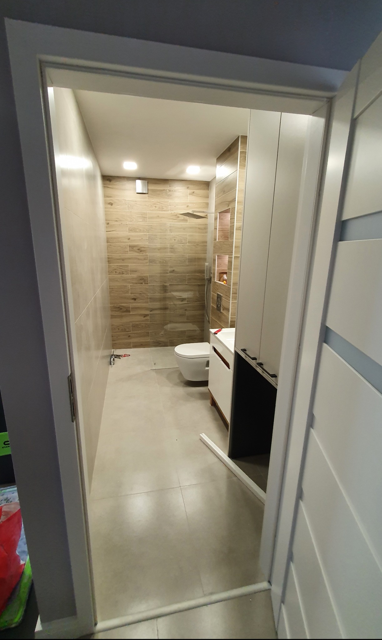 Nowoczesna łazienka z kabiną prysznicową typu walk-in, wykończona beżowymi płytkami imitującymi drewno, widoczna zza uchylonych białych drzwi.