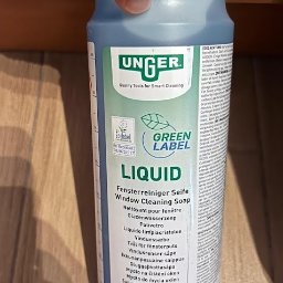 Green Hall Sp. z o.o. - Butelka płynu do mycia okien marki Unger, z zieloną etykietą i napisami w różnych językach, trzymana w dłoni na tle drewnianej powierzchni.