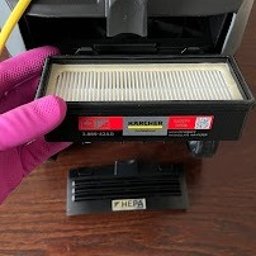 Green Hall Sp. z o.o. - Wymiana filtra HEPA w odkurzaczu Kärcher. Ręka w różowej rękawiczce trzyma filtr. Widoczny żółty kabel i fragment obudowy odkurzacza na drewnianym blacie.