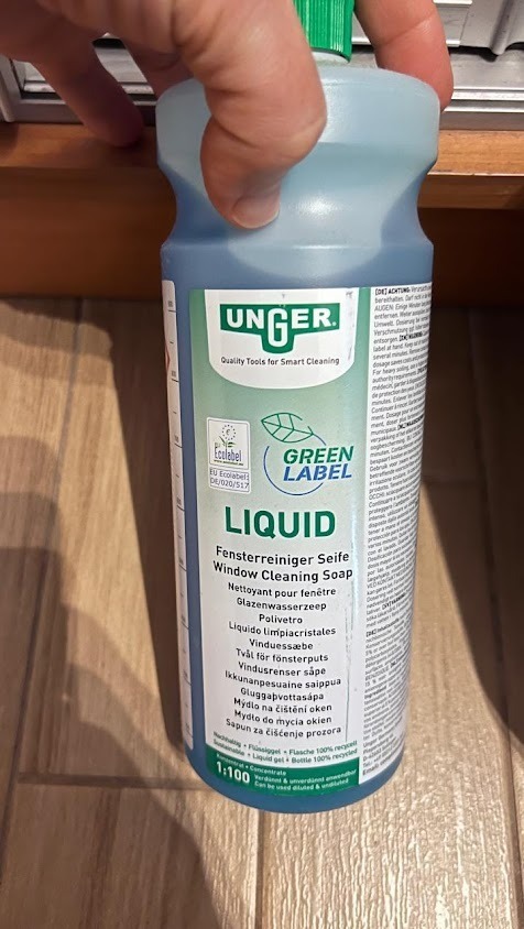 Butelka płynu do mycia okien marki Unger, z zieloną etykietą i napisami w różnych językach, trzymana w dłoni na tle drewnianej powierzchni.