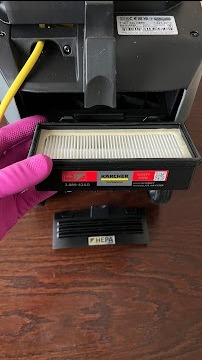 Wymiana filtra HEPA w odkurzaczu Kärcher. Ręka w różowej rękawiczce trzyma filtr. Widoczny żółty kabel i fragment obudowy odkurzacza na drewnianym blacie.