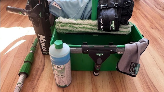 Zestaw profesjonalnych narzędzi do mycia okien marki Unger: kij teleskopowy Ninja, wiadro, ściągaczki, myjka, płyn do mycia i ściereczki z mikrofibry na drewnianej podłodze.