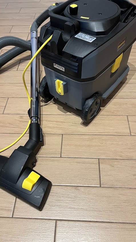 Profesjonalny odkurzacz Karcher z filtrem HEPA na drewnopodobnej podłodze. Widoczna ssawka i wąż. Sprzęt gotowy do użycia, sugerujący usługi porządkowe.