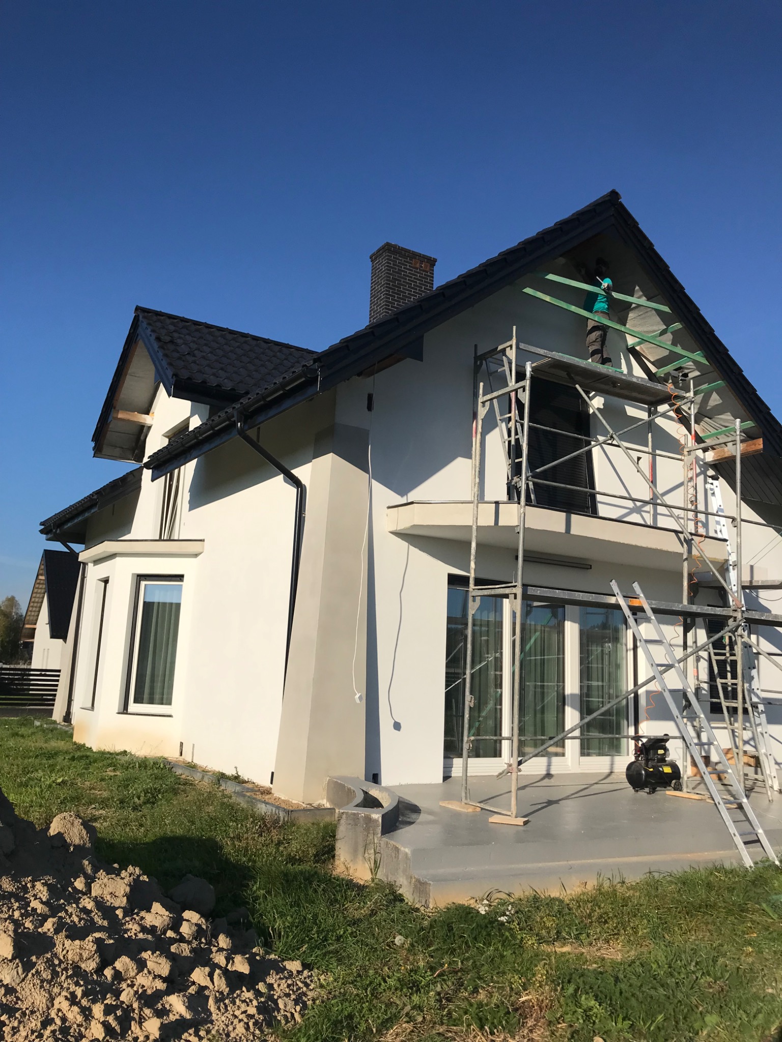 Elewacja domu jednorodzinnego w trakcie prac wykończeniowych. Widoczne rusztowania, pracownik montujący elementy podbitki dachowej. Świeżo otynkowana, biała fasada.