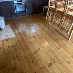 Perfect Clean - Wypolerowana, drewniana podłoga w kuchni z ciemnymi szafkami i ułożonymi krzesłami. Widoczne ślady po sprzątaniu, połyskująca powierzchnia.