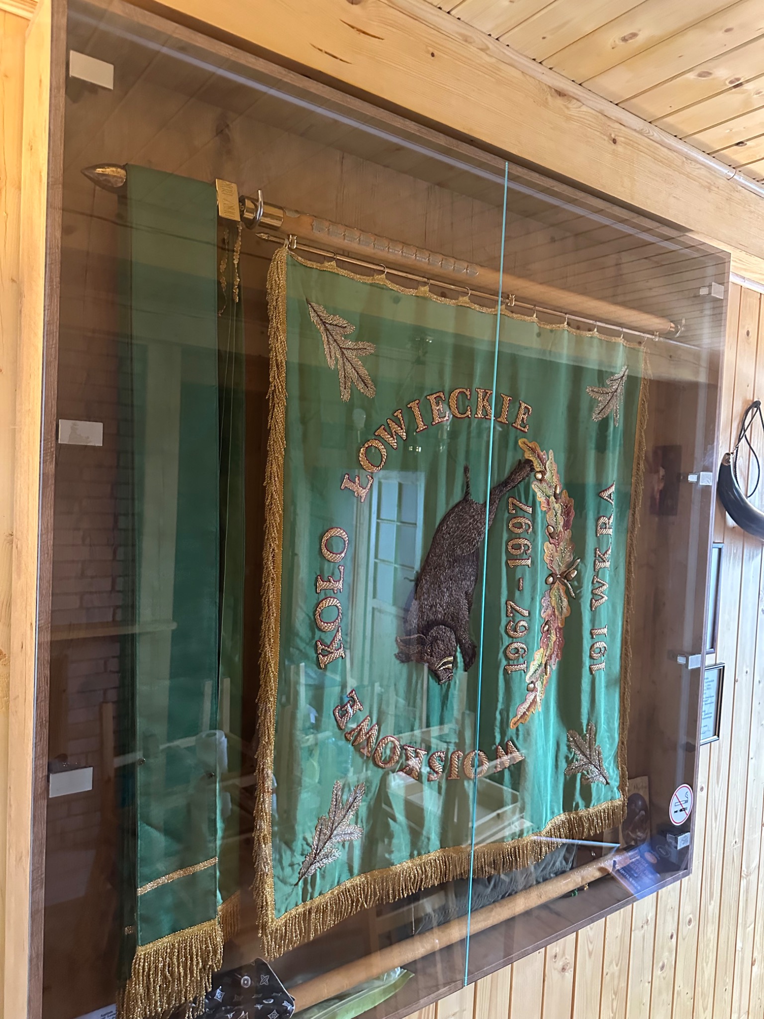 Zielony sztandar myśliwski w szklanej gablocie, z haftowanym dzikiem i napisem 'Koło Łowieckie'. Widoczne detale, takie jak frędzle i drewniane wykończenie.