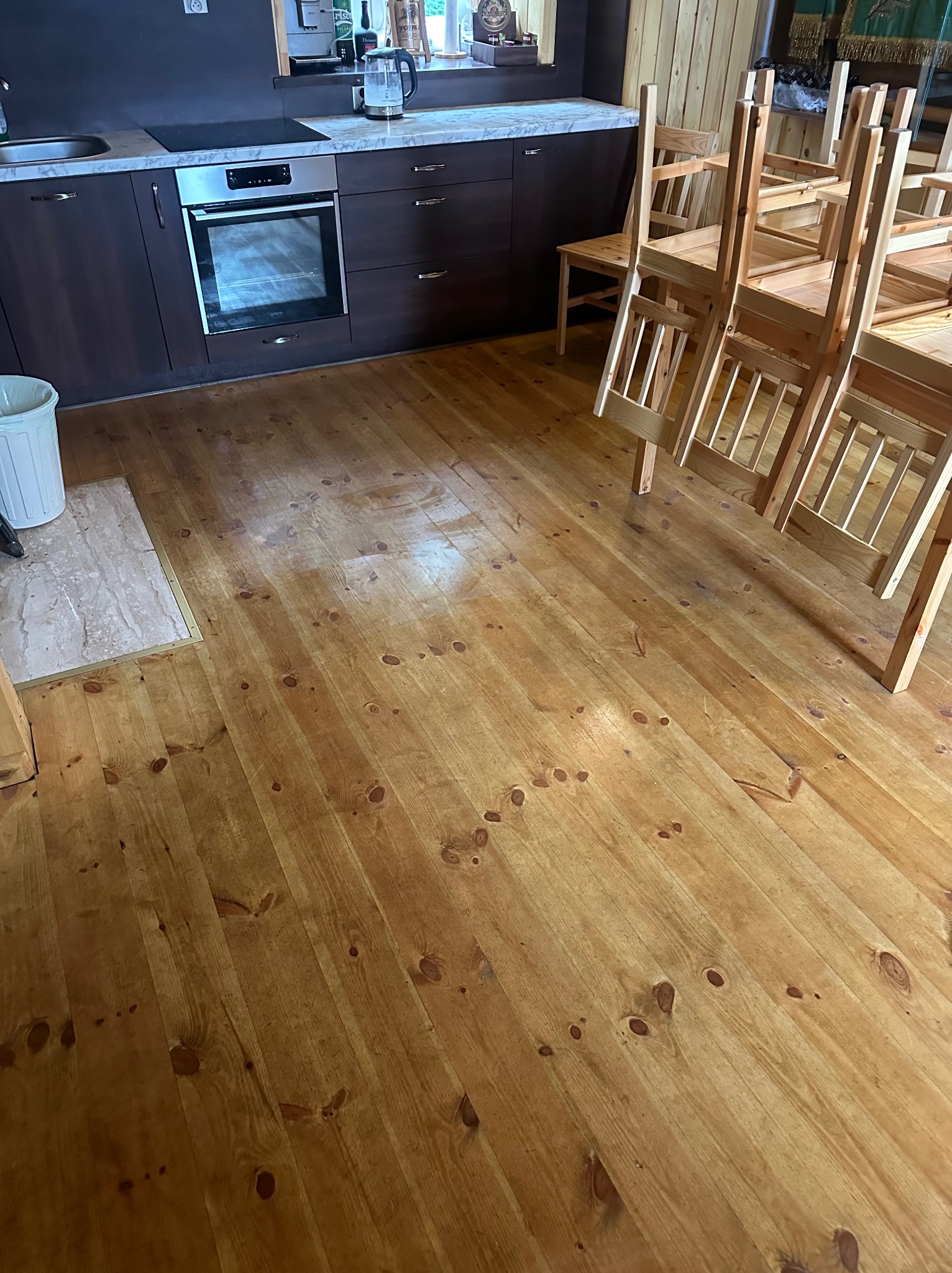 Wypolerowana, drewniana podłoga w kuchni z ciemnymi szafkami i ułożonymi krzesłami. Widoczne ślady po sprzątaniu, połyskująca powierzchnia.
