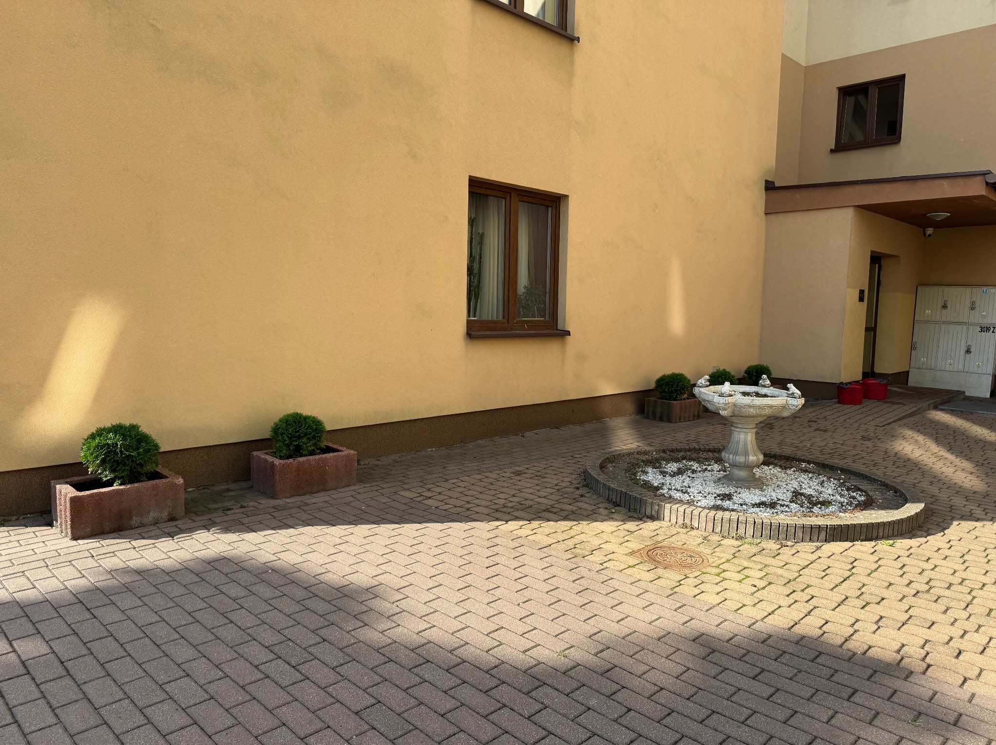 Dziedziniec budynku z fontanną, donicami i kostką brukową w słoneczny dzień. Widoczne okna i skrzynki pocztowe. Czysta przestrzeń, regularne wzory kostki.