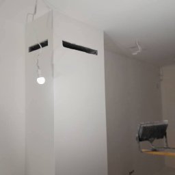 P&K BUD Ewa Kotlińska - Białe ściany po malowaniu z widocznymi otworami wentylacyjnymi w pionowym elemencie, oświetlone lampą budowlaną. Widoczne kable.