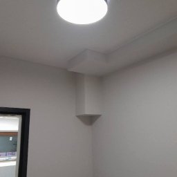 P&K BUD Ewa Kotlińska - Wnętrze po malowaniu: gładkie, jasne ściany, sufit z lampą i narożnikiem z półką. Widoczny fragment drzwi i okna. Minimalistyczny design.