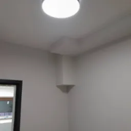 Wnętrze po malowaniu: gładkie, jasne ściany, sufit z lampą i narożnikiem z półką. Widoczny fragment drzwi i okna. Minimalistyczny design.