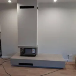 Nowoczesny kominek z obudową w odcieniach szarości, z widocznymi elementami instalacji elektrycznej i oświetleniem punktowym w minimalistycznym wnętrzu.