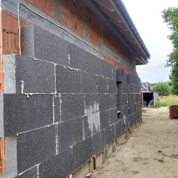 P&K BUD Ewa Kotlińska - Montaż grafitowych płyt termoizolacyjnych na ścianie budynku z czerwonej cegły. Widoczne spoiny wypełnione pianką montażową i fragment drewnianej konstrukcji dachu.