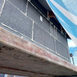 P&K BUD Ewa Kotlińska - Szara elewacja budynku w Kutnie, ocieplona styropianem grafitowym z wypełnionymi pianką szczelinami. Widoczne rusztowanie i fragment siatki ochronnej.